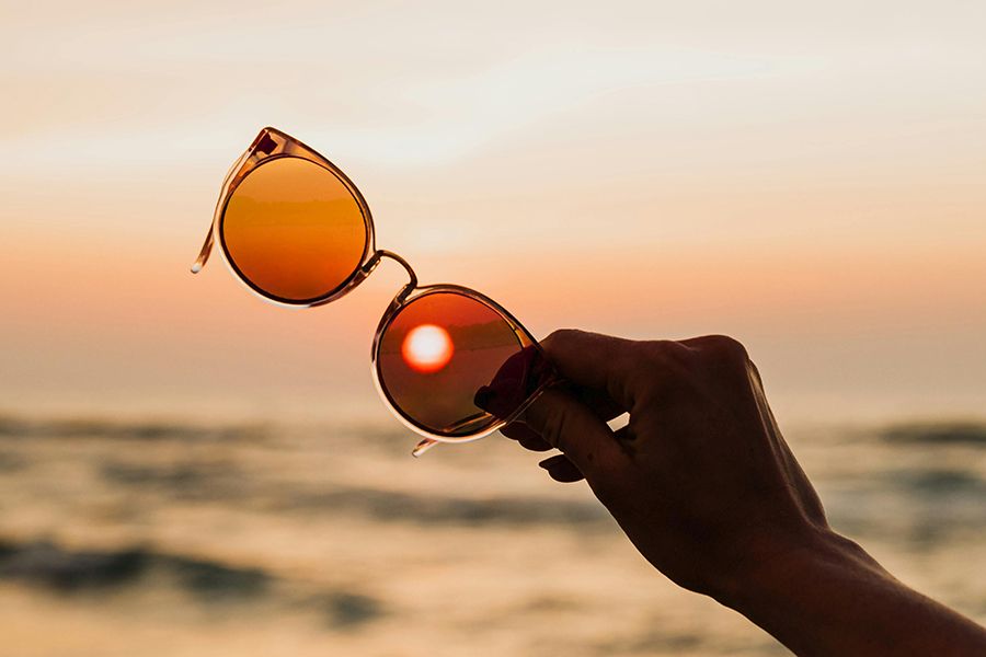 A hand raising sunglasses in front of the sea and the sun